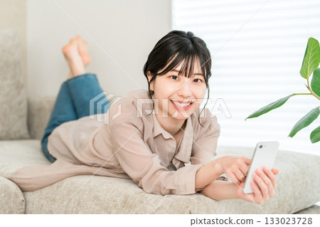 A young Asian woman lying on the sofa and relaxing while looking at her smartphone 133023728