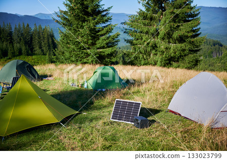 Tourist tent with solar panel and portable power station nearby in grassy field. Scene surrounded by rolling hills, tall pine trees under clear blue sky, showcasing eco-friendly camping setup. Tourist tent with solar panel and portable power station nearby in grassy field. Scene surrounded by rolling hills, tall pine trees under clear blue sky, showcasing eco-friendly camping setup. 133023799