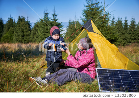 Young child and mother play with smartphone while charging with photovoltaic solar panel near tourist tent in summer. Integration of renewable energy in outdoor camping activities. Young child and mother play with smartphone while charging with photovoltaic solar panel near tourist tent in summer. Integration of renewable energy in outdoor camping activities. 133023800