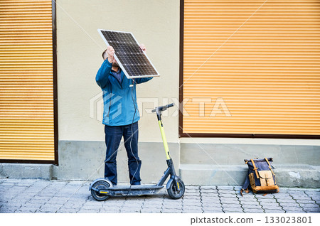 Male person holds photovoltaic solar panel, for charging electric scooter in urban settings. Integration of solar power as sustainable energy source, promoting eco-friendly urban transportation. 133023801