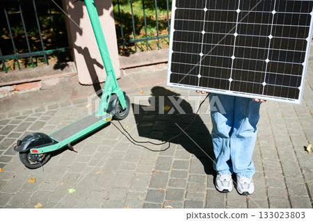 Woman using portable solar panel to charge scooter, highlighting integration of sustainable renewable energy. Green electric scooter connected to solar panel, emphasizing eco-friendly transportation. 133023803