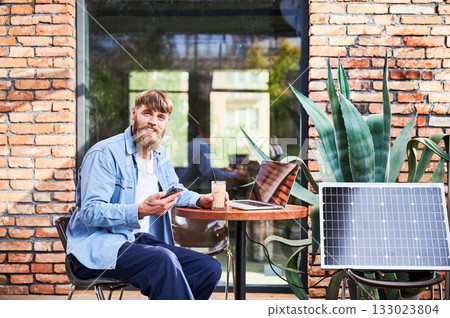 Man works on laptop and smartphone at outdoor table, with glass of iced coffee. Solar panel nearby, emphasizing sustainable, modern eco-friendly workspace that combines technology and sustainability. 133023804