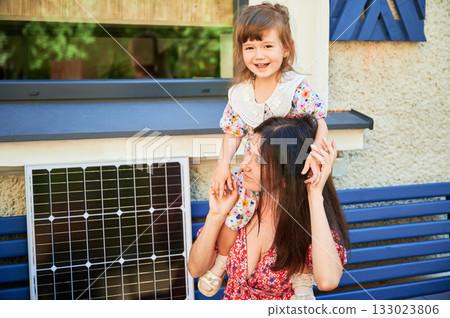 Mother sits on bench with her young daughter, both smiling joyfully. Solar panel placed beside them, symbolizing embrace of renewable energy and bright, sustainable future for next generation. Mother sits on bench with her young daughter, both smiling joyfully. Solar panel placed beside them, symbolizing embrace of renewable energy and bright, sustainable future for next generation. 133023806