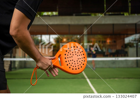 Player prepares to serve on padel court surrounded by greenery 133024407