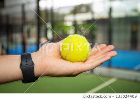 Hand holds a yellow padel ball on a green court 133024409