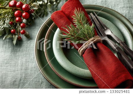 An elegant table setting of stacked green plates with a red cloth napkin, surrounded by festive greenery and oak branches, creates a warm and cozy atmosphere for the Christmas table. An elegant table setting of stacked green plates with a red cloth napkin, surrounded by festive greenery and oak branches, creates a warm and cozy atmosphere for the Christmas table. 133025481