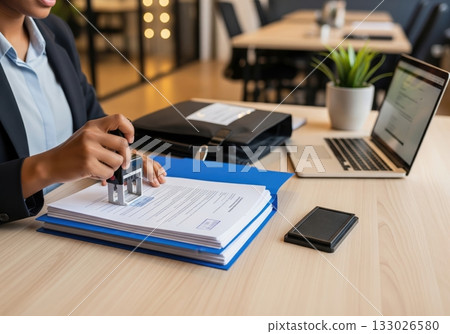 In an office setting, a businesswoman stamps a document from a file, a process integral to business compliance, and alongside a laptop and briefcase 133026580