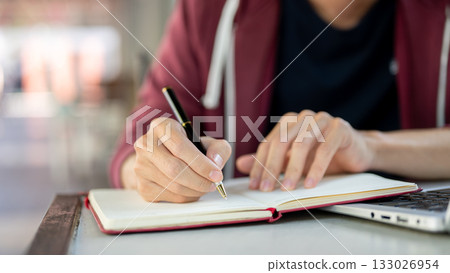 A hand holding ballpoint pen writing in red cover notebook while sitting with a laptop at the table. A hand holding ballpoint pen writing in red cover notebook while sitting with a laptop at the table. 133026954