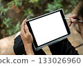 Close up of a hand holding white screen tablet and stylus while sitting cross legged near plant bed. 133026960