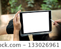 Close up of a hand holding blank screen tablet and stylus while sitting cross legged near plant bed. 133026961