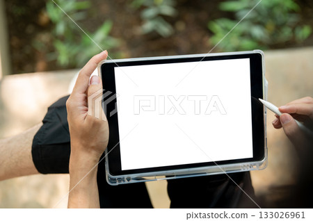 Close up of a hand holding blank screen tablet and stylus while sitting cross legged near plant bed. Close up of a hand holding blank screen tablet and stylus while sitting cross legged near plant bed. 133026961