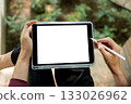 Top view of a hand holding white screen tablet and stylus pen while sitting cross legged near plant bed. 133026962