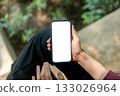 Close up of hand holding and hovering over white screen phone while sitting cross leg near plant bed 133026964