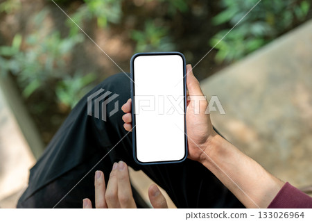 Close up of hand holding and hovering over white screen phone while sitting cross leg near plant bed 133026964