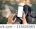 Top view of hand holding and hovering over white screen phone while sitting cross legged in the park 133026965