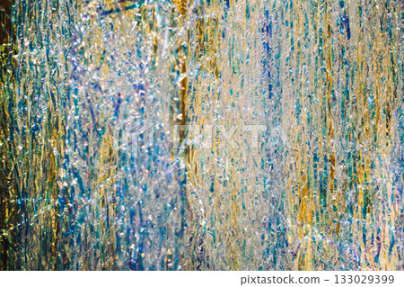 Close-up of iridescent tinsel strands shimmering in layered metallic colors under bright light. Immersive event backdrops, sensory holiday displays, touchable decor, visual styling trends Close-up of iridescent tinsel strands shimmering in layered metallic colors under bright light. Immersive event backdrops, sensory holiday displays, touchable decor, visual styling trends 133029399