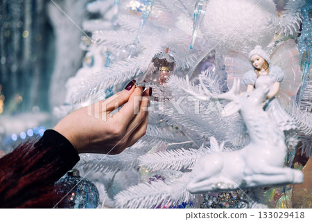 Hand placing a delicate bell ornament on a white Christmas tree adorned with fairy and reindeer decorations. Holiday bonding moments, personal touch in decor, festive rituals, seasonal creativity Hand placing a delicate bell ornament on a white Christmas tree adorned with fairy and reindeer decorations. Holiday bonding moments, personal touch in decor, festive rituals, seasonal creativity 133029418