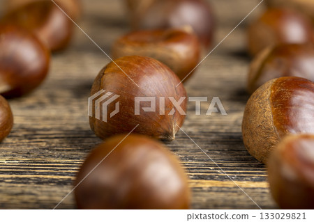 a hard edible chestnut fruit in a brown shell on a wooden table, a group of raw fruits of chestnut close up a hard edible chestnut fruit in a brown shell on a wooden table, a group of raw fruits of chestnut close up 133029821
