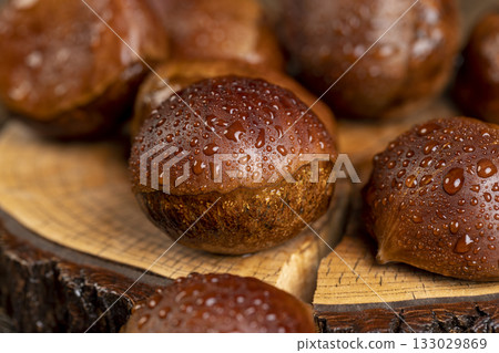 a hard edible chestnut fruit in a brown shell covered with water droplets on a wooden table, a group of wet and fresh chestnut with a brown hard shell 133029869