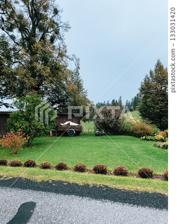 Lush green grass enveloping wooden shed and trailer, with trees displaying vibrant fall colors under cloudy sky in serene Austrian countryside. Wooden shed nestled among lush green grass in Austrian 133031420