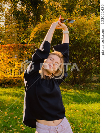 Happy young blonde woman wearing black sweater and pink pants, holding sunglasses while stretching in park on sunny autumn day, surrounded by vibrant yellow leaves and trees. Woman in autumn park Happy young blonde woman wearing black sweater and pink pants, holding sunglasses while stretching in park on sunny autumn day, surrounded by vibrant yellow leaves and trees. Woman in autumn park 133031426