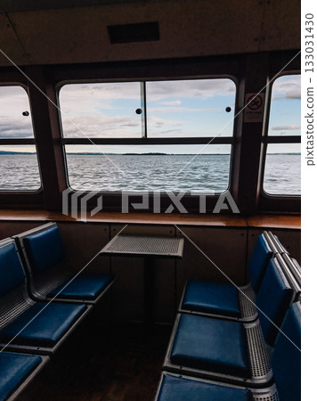 Empty passenger seats and small table inside ferry boat navigating on Lake with view on water and cloudy sky through large windows. Vacant ferry seats, small table, large windows revealing Lake waters 133031430