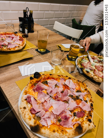 Round Neapolitan pizza with cooked ham and mushrooms, served on white plate placed on wooden table set with yellow placemats, glasses and beer. Customers are having dinner. Meeting friends for pizza 133031448
