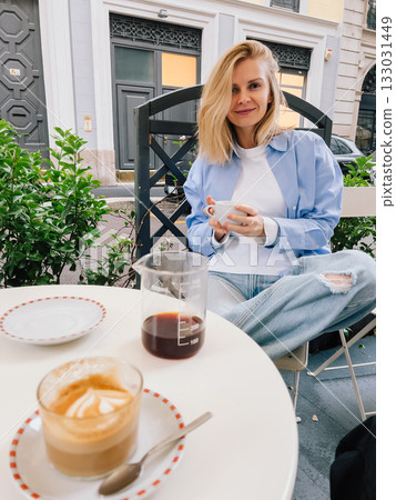 Young woman sitting at table in bustling street cafe, savoring rich cup of coffee and creamy cappuccino while dressed in casual attire and enjoying vibrant urban atmosphere. Woman sipping cappuccino 133031449