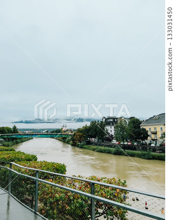 River winding through historic city, revealing colorful cityscape, arching bridge, alpine landscape with mountain under cloudy skies. River flowing through city on cloudy day with colorful bridge River winding through historic city, revealing colorful cityscape, arching bridge, alpine landscape with mountain under cloudy skies. River flowing through city on cloudy day with colorful bridge 133031450