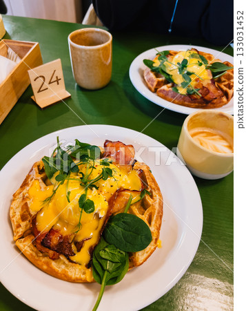 Delicious savory waffles topped with poached eggs, crispy bacon, fresh greens, and creamy hollandaise sauce are being served on white plate in cafe, creating gourmet brunch experience. Gourmet brunch Delicious savory waffles topped with poached eggs, crispy bacon, fresh greens, and creamy hollandaise sauce are being served on white plate in cafe, creating gourmet brunch experience. Gourmet brunch 133031452