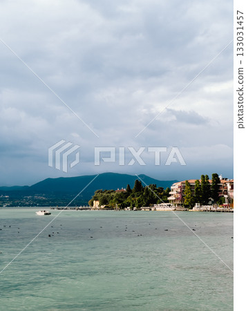 Motorboat gliding across tranquil waters of Lake Garda, surrounded by charming hotels and restaurants along coast, with majestic mountains rising under cloudy sky. Motorboat cruising Lake Garda waters 133031457