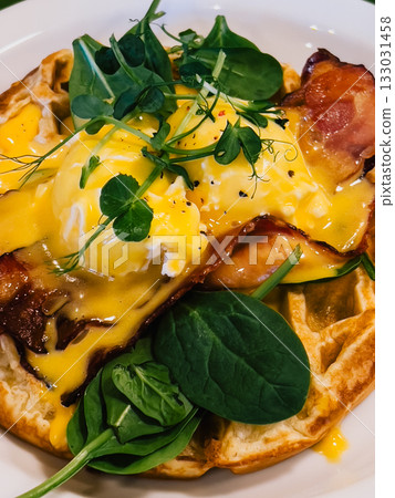 Close up of savory waffle topped with poached eggs, crispy bacon, creamy hollandaise, fresh spinach, and delicate pea sprouts, creating appetizing brunch dish. Gourmet brunch plating. Waffle, eggs 133031458
