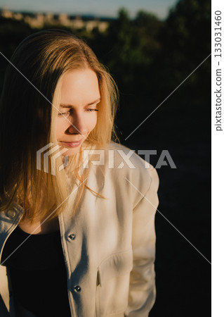 Young blonde woman wearing light jacket is smiling and looking down, enjoying warm sunlight during golden hour, with blurred background of trees and cityscape. Woman basking in golden hour sunlight Young blonde woman wearing light jacket is smiling and looking down, enjoying warm sunlight during golden hour, with blurred background of trees and cityscape. Woman basking in golden hour sunlight 133031460