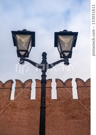 Vintage black forged street lamp against an old red brick fortress wall with battlements. Urban background for the concept of history, security, and city street illumination. 133031611