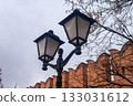 Vintage black forged street lamp against an old red brick fortress wall with battlements. Urban background for the concept of history, security, and city street illumination. 133031612