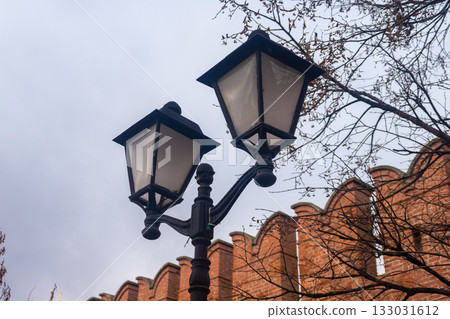 Vintage black forged street lamp against an old red brick fortress wall with battlements. Urban background for the concept of history, security, and city street illumination. Vintage black forged street lamp against an old red brick fortress wall with battlements. Urban background for the concept of history, security, and city street illumination. 133031612