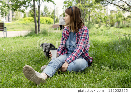 Young woman sitting on grass with coffee cup and small mixed breed gray fluffy senior dog bedlington terrier whippet enjoying a sunny day in a park with greenery 133032255