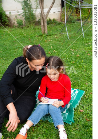 Mother and daughter outdoors, drawing. Bonding and creativity in a lush green yard. A cozy, intimate moment of learning and growth. 133033081