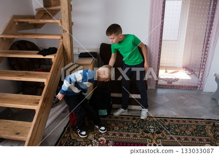 Two boys are near the stairs in a home, one is wearing a blue and white sweater and black jeans and the other is in a green shirt and jeans 133033087