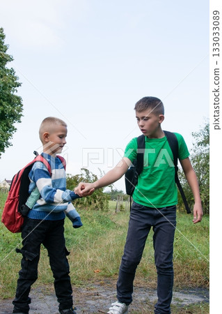 Two young boys, returning from school, walk on a path in the grass. One boy is helping his younger friend. Both are wearing backpacks. 133033089