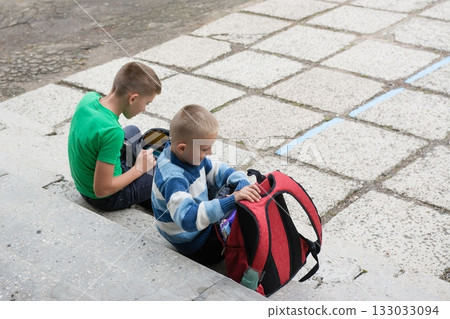 Two boys getting ready on stone steps before school. One packs a red backpack, the other uses an electronic device in his black bag. Two boys getting ready on stone steps before school. One packs a red backpack, the other uses an electronic device in his black bag. 133033094