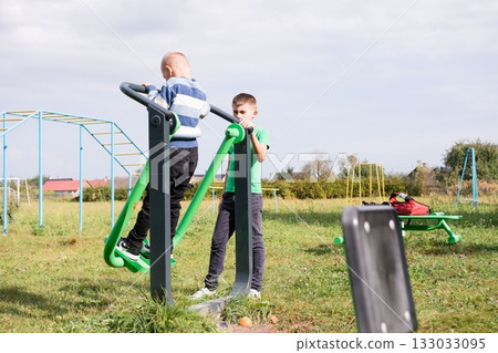 Two boys are playing on outdoor gym equipment at a local park on a sunny day. There are other play structures in the background. Two boys are playing on outdoor gym equipment at a local park on a sunny day. There are other play structures in the background. 133033095