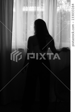 Young woman in soft light, standing at window looking outside, contemplating future, feeling hope, privacy and introspection, black and white portrait 133033164