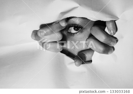 The eye of a young girl peering through a hole in a piece of white paper, her hands tearing at the edges, conveying feelings of curiosity and fear 133033168