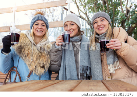 Portrait three pretty young adult woman friends enjoy laughing drinking hot mulled wine at Christmas market outdoors cold winter day. People having fun celebrate new year time street cafe outside 133033173