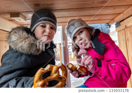 Portrait two cute little funny children enjoy eating german pretzel sitting under wooden table at cafe outdoors cold winter day. Cheerful kids friends playing outside together frosty weather 133033176