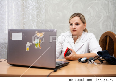 Physician engages in telehealth, wearing a white coat while consulting online with blood pressure cuff and stethoscope nearby. Physician engages in telehealth, wearing a white coat while consulting online with blood pressure cuff and stethoscope nearby. 133033269