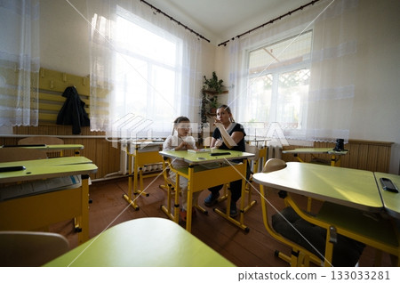 Teacher and young student in classroom. The pair sit at desks under bright windows in the well lit educational space. 133033281