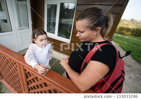 Mother and daughter at a wooden railing. A young girl holds an object while conversing with her mother near a building. Mother and daughter at a wooden railing. A young girl holds an object while conversing with her mother near a building. 133033286