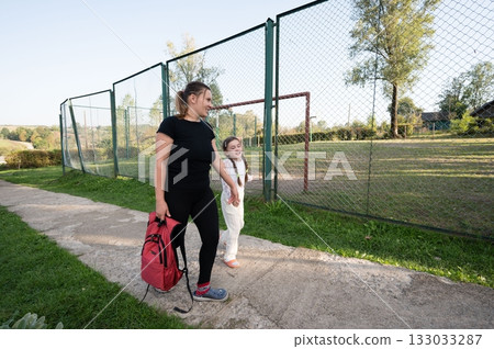 Mother and daughter stroll along a path beside a fenced field, sharing a moment of connection on a sunny day. Mother and daughter stroll along a path beside a fenced field, sharing a moment of connection on a sunny day. 133033287